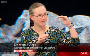 A woman on a red sofa. She is wearing glasses and gesturing. In the background is a large screen showing a grey lunar landscape.
